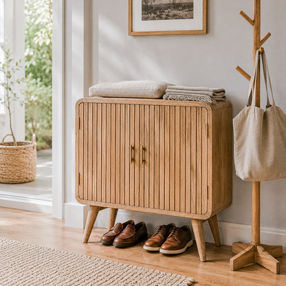 A light natural wood entryway cabinet with vertical slats and tapered legs, styled in a bright room with folded blankets on top, shoes underneath, a coat rack with a tote bag, and a window in the background.