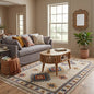 A styled living room featuring a large cream-colored area rug with a colorful geometric pattern and fringe, styled with a gray sofa, wooden coffee table, and potted plant.